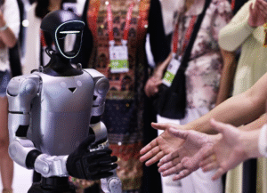 Humanoid robot with a glossy gray body and glowing blue visor stands in front of a crowd, people reaching out to greet it.